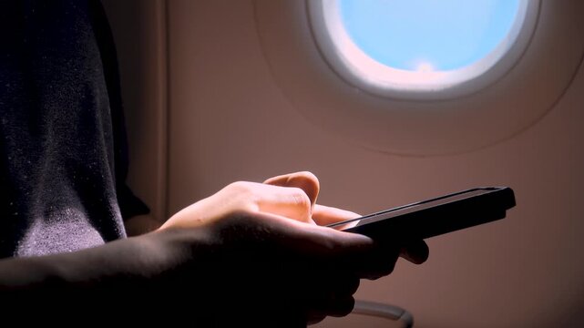 Close up unrecognizable passenger using smartphone during airplane flight near window. Typing messages scrolling social media playing games reading content on mobile device in air travel cabin