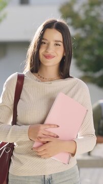 Portrait of a beautiful young woman smiling at the camera while standing outdoors on the university campus, holding a pink notebook. Vertical