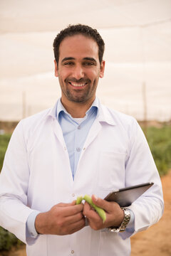 Professional Middle Eastern male agricultural scientist wearing white lab coat smiles while holding digital tablet and inspecting fresh organic