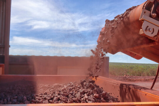 Hematite iron ore dropping off conveyor. Mine, mining and processing. Dust rising as crushed red rock are discharges into a hopper or feeder. Copy space in sky