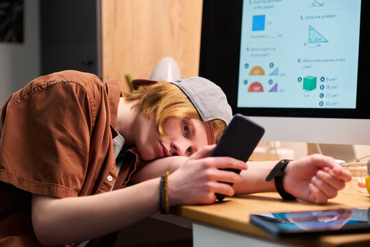 Teenage boy using smartphone while resting head on desk beside online math lesson, showing study distraction and screen addiction. Useful for education, teen digital habits marketing
