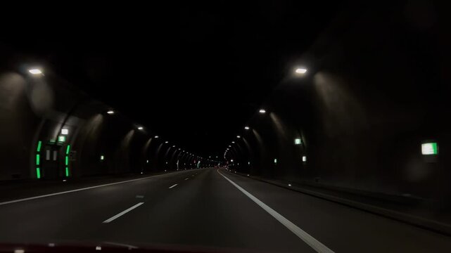 Driving through the modern, dimly lit tunnel on the expressway at night, captured from a car front window view moving forward along the illuminated empty road asphalt.