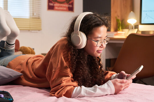 Asian teenage girl using smartphone while wearing headphones at home, engaging in digital entertainment and mobile communication. Useful for teen screen time or online media campaigns