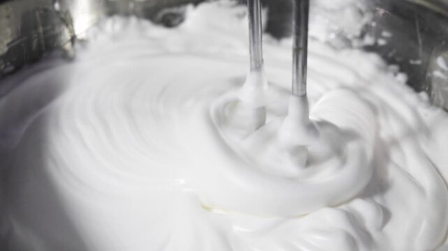 Close-up of an electric mixer beating egg whites and sugar into a smooth, glossy meringue. Food preparation for baking pastries and cakes
