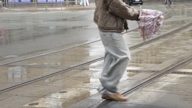 Pedestrians walking across wet tram tracks on a city street during rainy weather, showcasing urban transit infrastructure and daily commute