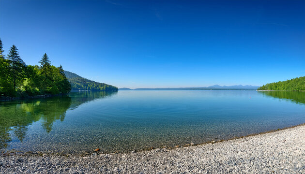 A Serene Lakeside Scene Featuring A Pebble Beach Lush Greenery And A Vast Expanse Of Calm Water Under A Clear Blue Sky