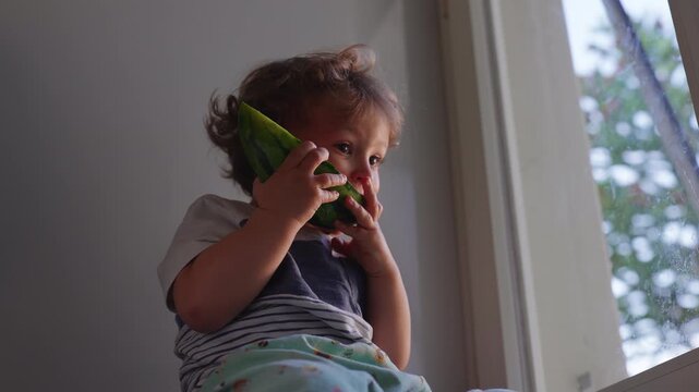 Toddler eating watermelon while sitting by open window at home, healthy childhood nutrition moment highlighting curiosity, wellbeing, independence and sensory exploration