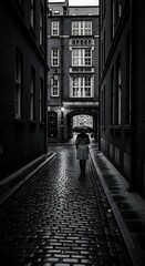 Fototapeta premium Lonely person walking through a dark cobblestone alleyway in a city.