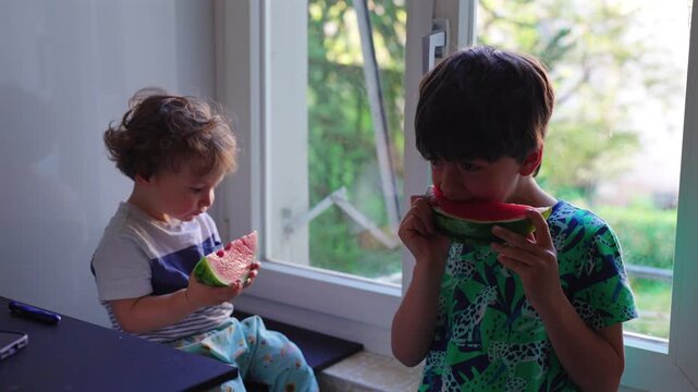 Siblings eating watermelon together by window at home, healthy childhood nutrition scene highlighting family connection, wellbeing, freshness and shared moments