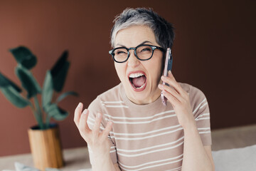 Naklejka na ściany i meble Frustrated middle-aged woman yelling on phone during a heated call at home, expressive emotion and stress captured in candid portrait