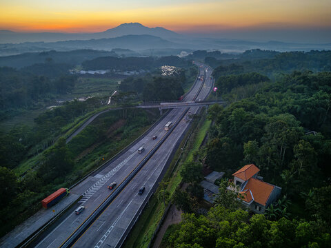 Aerial view of the Jalan Tol Bawen - Salatiga highway winding through lush green hills and forests at sunset with distant mountains in Tengaran, Central Java, Indonesia.