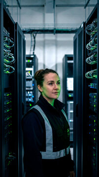 Serious female technician in safety vest standing between server racks in data center