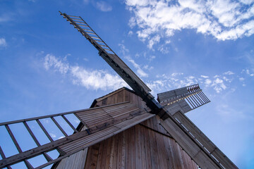 Traditional wooden post mill windmill viewed from below against a blue sky with white clouds. Historic rural architecture and cultural heritage landmark showcasing traditional grain milling technology © MAbakanowicz