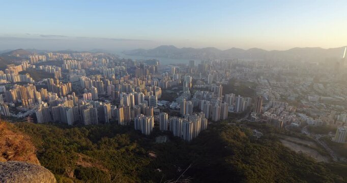 Cityscape of Kowloon peninsula with dense clusters of residential high-rises seen from Lion Rock Country Park. Panning shot in warm evening sunlight reveals vast urban sprawl of Hong Kong
