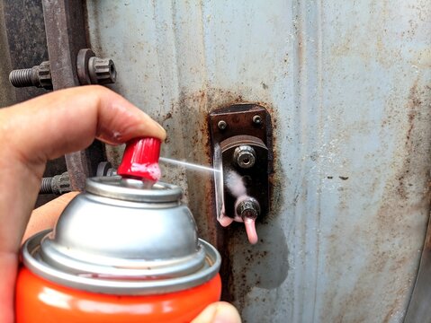 Close-up of a hand applying rust penetrant spray to heavily corroded bolt and nut components, demonstrating maintenance procedures, metal restoration, and industrial repair work