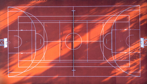 Aerial view of a red basketball court with white markings and long tree shadows cast across the surface in Suchacz, Warmian-Masurian Voiv, Poland.
