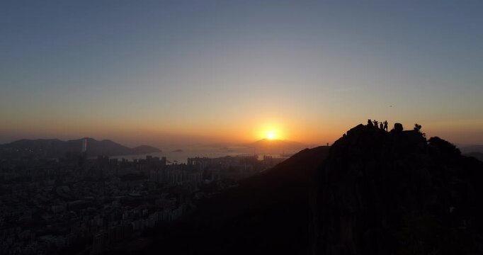 Captivating sunset moment in Hong Kong, with silhouetted Lion Head rock and tiny hikers figures atop at right. Bright yellow sun touches horizon, casting dramatic light