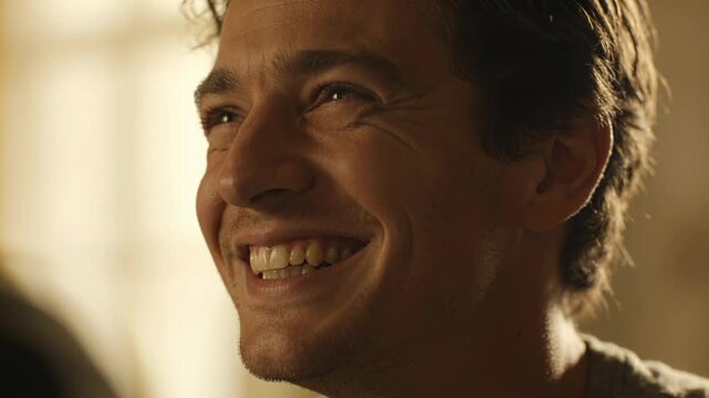 Smiling man reacting to off-cam pal joke at home, light tee, warm backlit window, blurred shoulder