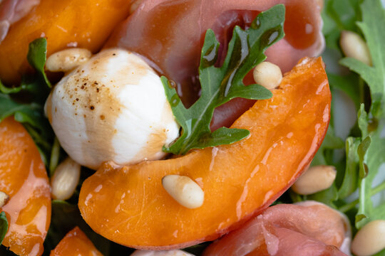 Macro close-up of a fresh summer salad featuring juicy apricot slices, creamy mozzarella pearls, delicate prosciutto, peppery arugula, and toasted pine nuts.