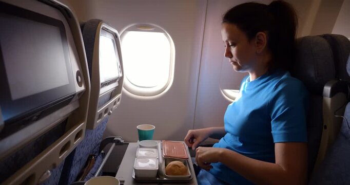 Passenger woman opens foil meal box during flight, glances inside with puzzled surprise, then pushes it away in visible refusal. Economy class in-flight service, side view. Food appears unappetizing