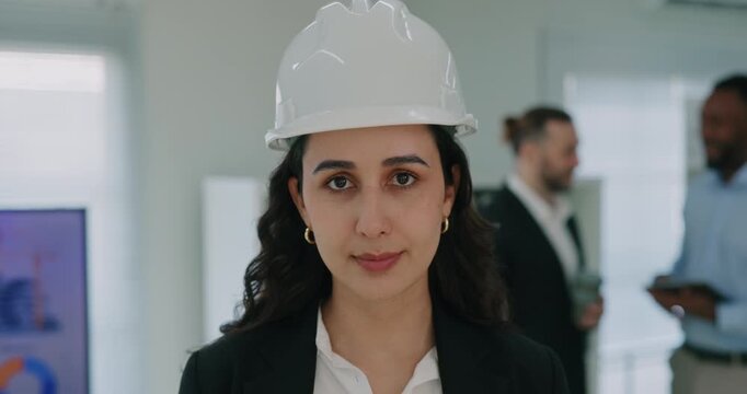 Portrait of brunette Latin American lady in white helmet stands in building company office. Hispanic woman contractor expresses professionalism at presentation