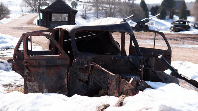 Burned rusted car shell with open front doors sits in melting snow at rural scrapyard, showing exposed chassis, missing windows, charred metal, and quiet
