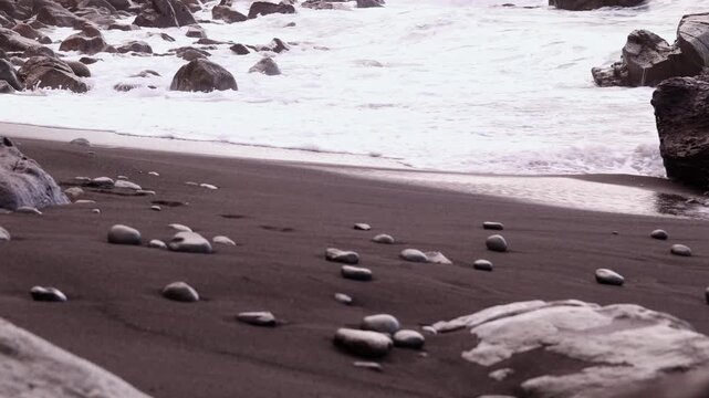 Back view of young woman in gray sweater and white pants walking barefoot along black volcanic sand beach in Tenerife toward crashing Atlantic ocean waves, stones visible on sand. Cinematic travel fas