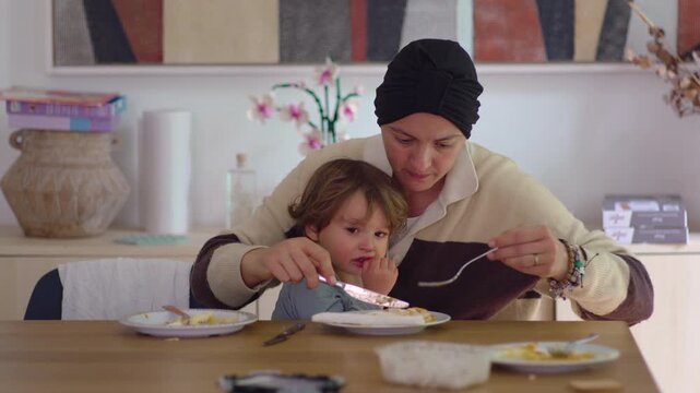 Mother eating meal while toddler rests on her lap at dining table in family home during cancer recovery, highlighting caregiving, resilience, emotional support and family connection