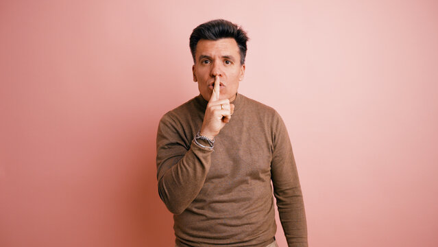 Man making a shushing gesture, requesting silence or discretion with his finger on his lips on a pink background