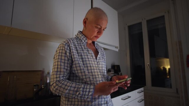 Female cancer patient checking smartphone at night in home kitchen during recovery, highlighting communication, resilience, support network, connection and modern daily life