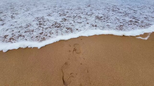Slow motion tracking backward as sea water and white foam wash over footprints on sandy beach.