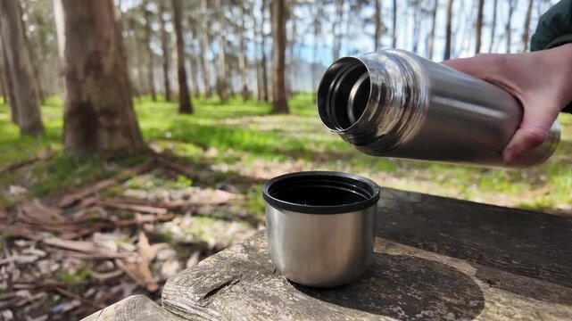 Pouring hot tea from steel thermos into cup a rustic wooden picnic table in forest, slow motion