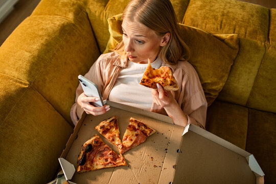 Young woman eating pizza and using smartphone on sofa at home