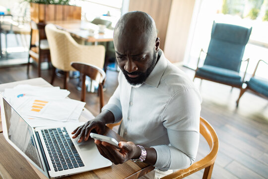 Worried businessman checking smartphone by laptop in cafe