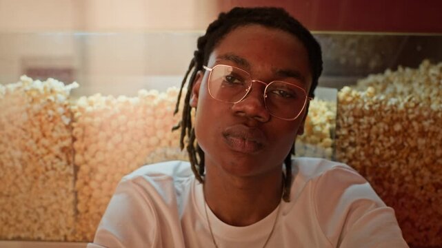 Zoom out slowmo portrait of African American young adult man with glasses looking at camera in front of popcorn display. Clip is ideal for cinema promotions and youth entertainment marketing campaigns