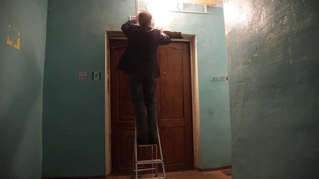 Man on ladder inspecting or replacing a light bulb above front door entrance.