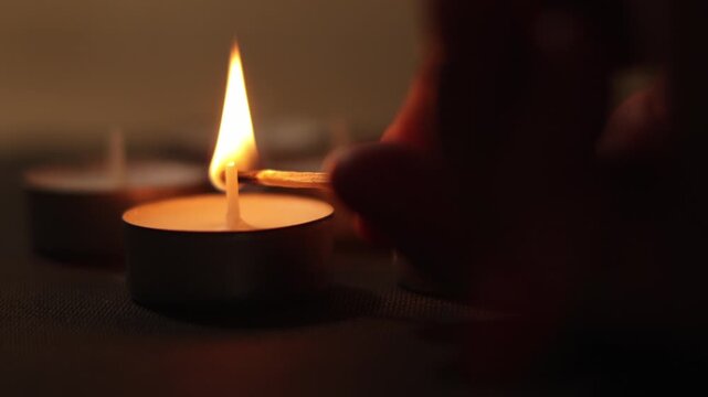 A close-up shot of a hand lighting multiple tea light candles one by one using a wooden matchstick. Romantic warm light atmosphere with beautiful shallow depth of field and soft bokeh on the backgroun