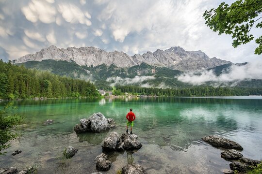 Young man standing on a rock on the shore, Eibsee lake in front of Zugspitze massif with Zugspitze, dramatic Mammaten clouds, Wetterstein range, near Grainau, Upper Bavaria, Bavaria, Germany