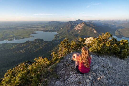 Young woman sitting at the summit, view from Herzogstand to Lake Kochel, Jochberg and Lake Walchen, Alpine foothills, Alps, Upper Bavaria, Bavaria, Germany