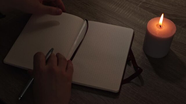 A peaceful and atmospheric video capturing the end of a creative process or evening routine. A person finishes writing in a checkered notebook, closes it, secures it with an elastic band, and places a