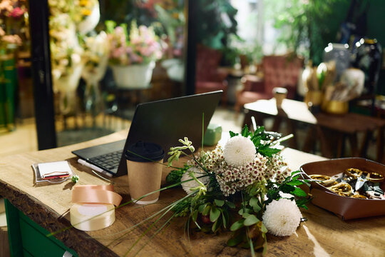 Close up of florist workspace showing laptop, bouquet, ribbon, coffee cup, shears on table, supporting flower shop operations. Ideal for small business, ecommerce, creative work themes