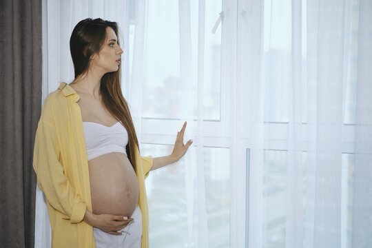 A pregnant woman stands by a window in a contemporary apartment, wearing a white top and loose yellow shirt. She gently touches her belly and gazes thoughtfully outside, basking in the natural light