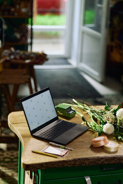 Vertical shot of laptop displaying blank screen on worktable beside flowers and stationery, supporting small business content creation, online shop branding, product marketing