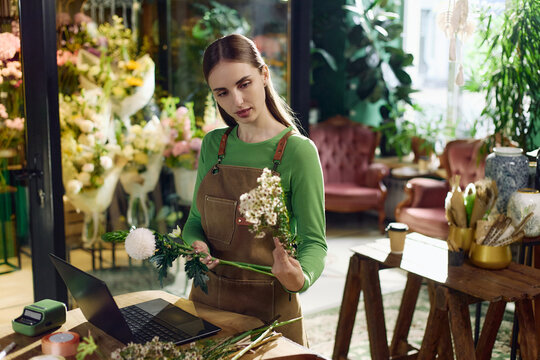 Young adult Caucasian woman florist arranging flowers while checking laptop in flower shop, managing small business workflow. Ideal for retail, entrepreneurship, ecommerce, vertical