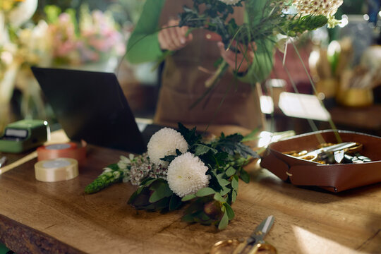Close up of florist arranging bouquet on worktable, using laptop for order management in flower shop. Ideal for small business marketing, retail branding, ecommerce content