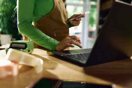 Unrecognizable woman managing online orders on laptop while holding smartphone at retail counter, processing small business sales and digital payments for ecommerce marketing