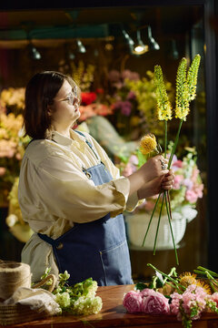 Vertical shot of young adult florist arranging fresh flowers in shop, selecting stems for bouquet design. Ideal for small business marketing, retail branding, craft entrepreneurship