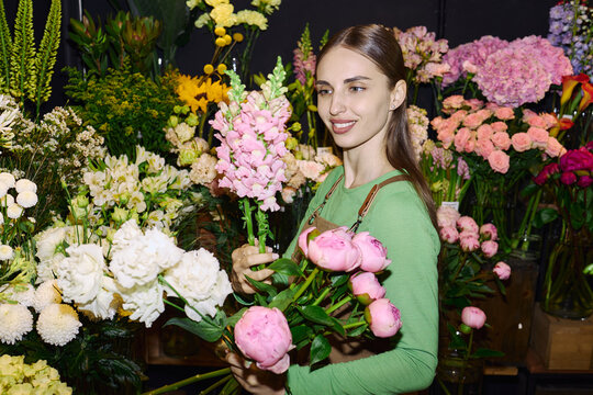 Young adult Caucasian woman selecting fresh flowers and arranging bouquet in florist shop, showing retail floral business and gift shopping concept. For ecommerce, local store marketing, wide shot