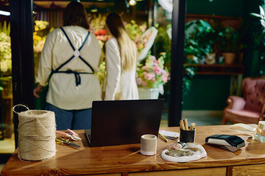 Wide shot of female florist and woman discussing bouquet order in flower shop, with laptop on counter supporting retail workflow. Ideal for small business, ecommerce, service marketing
