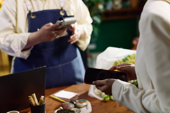 Close up of young adult woman holding smartphone while cashier processing contactless payment. Illustrating mobile checkout, retail technology, cashless commerce